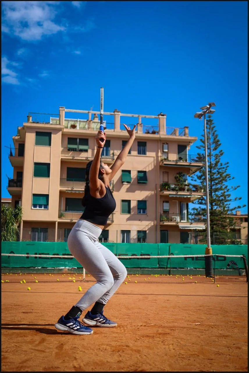 A player at full stretch on the ball toss, serving on an outdoor clay court