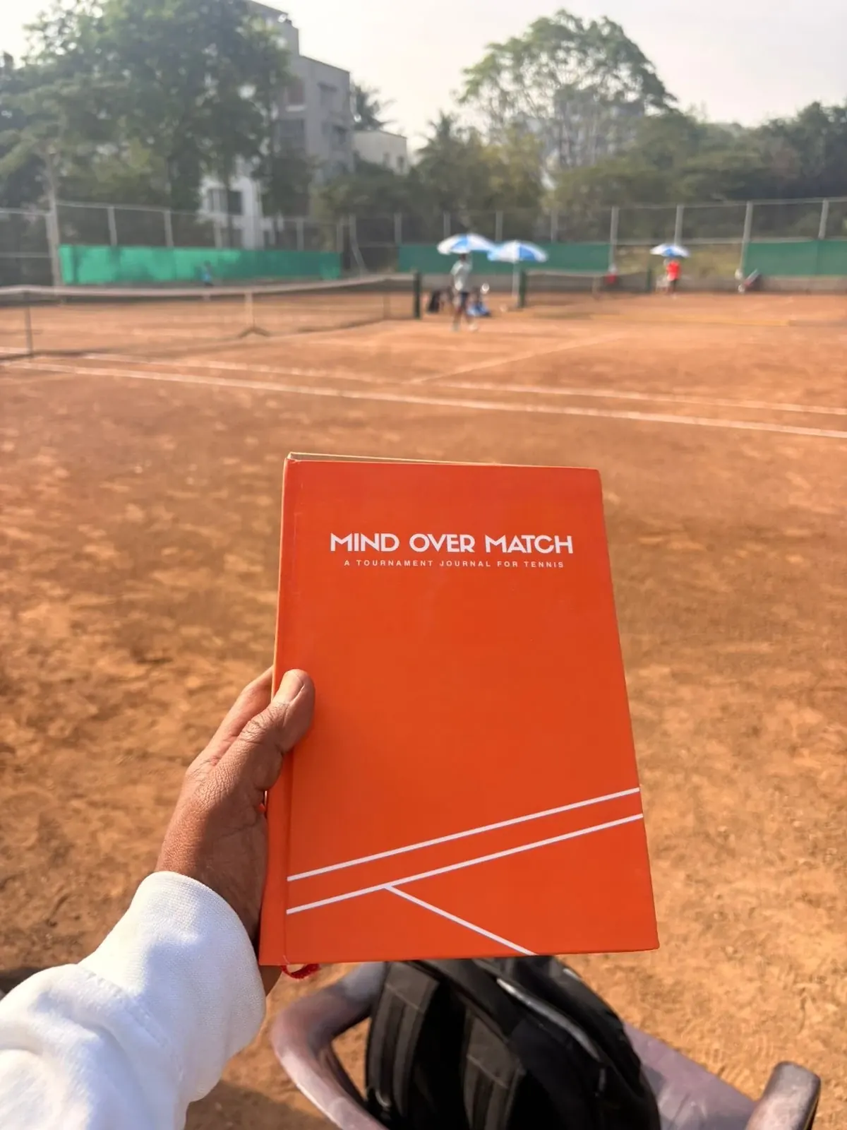 A player holding the Tovio Tournament Journal at the side of a clay court, a match going on behind